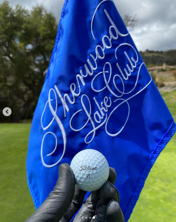 Brandon Thomas Lee hält einen Golfball vor einer blauen Flagge mit dem Logo des Sherwood Country Club, gepostet im März 2021 | Quelle: Instagram/brandonthomaslee