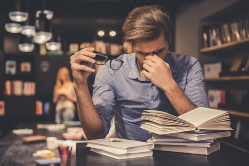 Der müde junge Mann hält Brillen und massiert seine Nasenbrücke, während er in der modernen Bibliothek hart arbeitet. I Quelle: Shutterstock