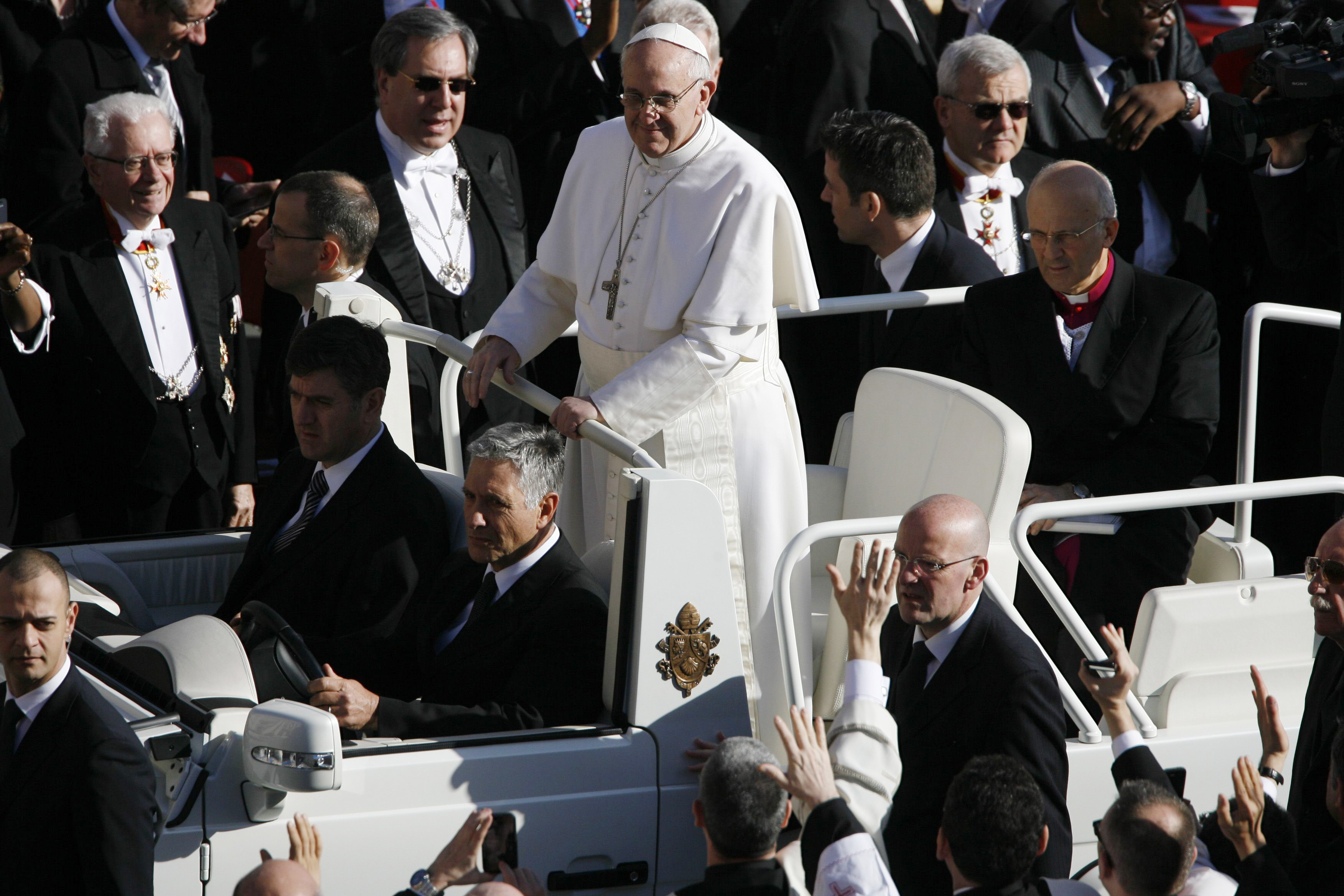 Papst Franziskus steht im Papamobil, umgeben von einem Meer von Würdenträgern und winkenden Händen, ganz in Weiß gekleidet und mit einem silbernen Kreuz über der Brust