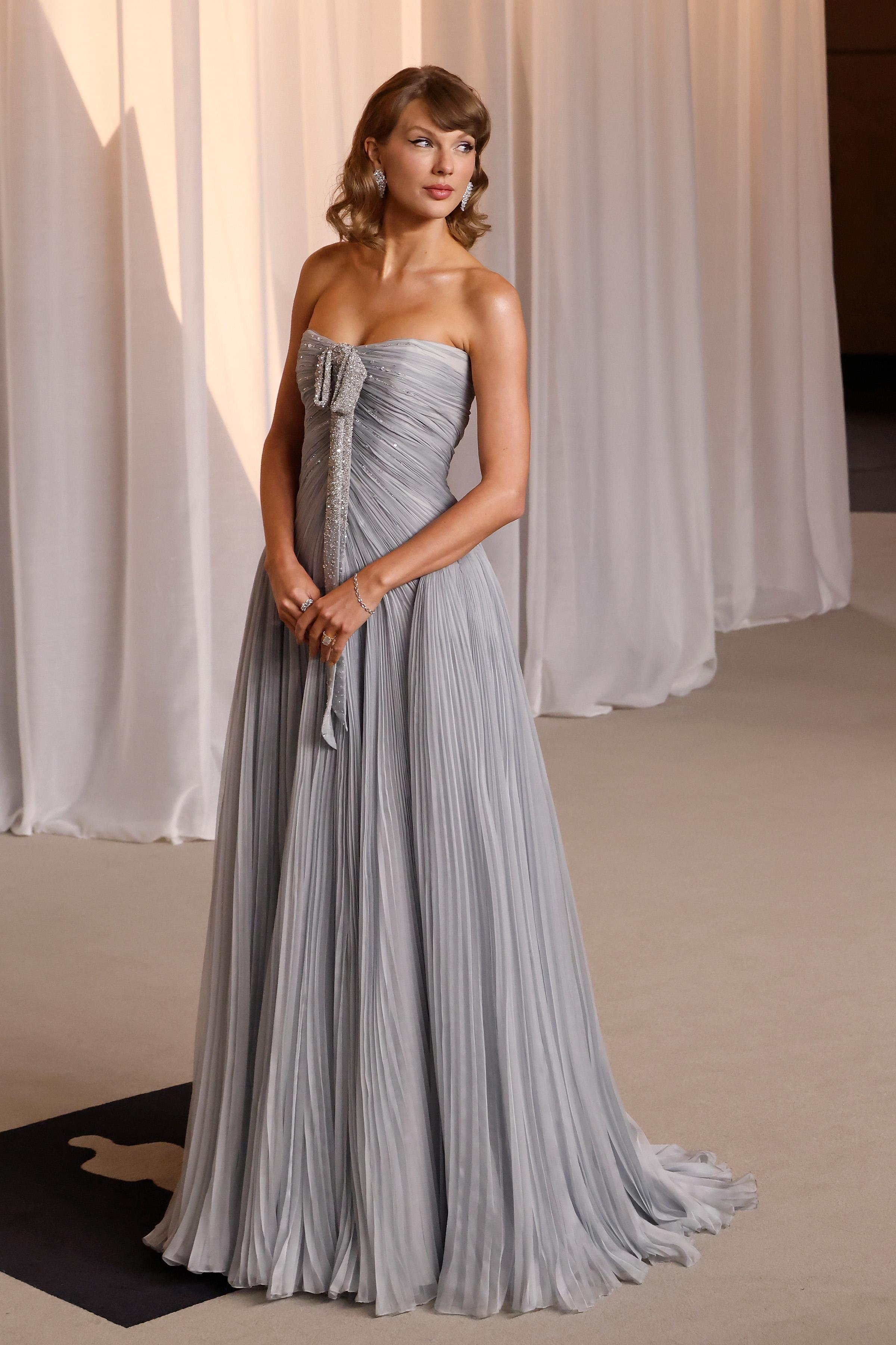 Taylor Swift strahlt in einem trägerlosen, taubengrauen Abendkleid, das durch zarte Plissees am Oberteil und am weit schwingenden Rock akzentuiert wird, eine himmlische Eleganz aus. Ein schimmerndes, mit Kristallen verziertes Schleifendetail fällt anmutig über die Vorderseite, ergänzt durch funkelnde Diamant-Ohrhänger und sanfte Vintage-Wellen. Bereit, als musikalischer Höhepunkt des Abends die Bühne zu betreten, versprüht die Grammy-Gewinnerin puren, zeitlosen Glamour.