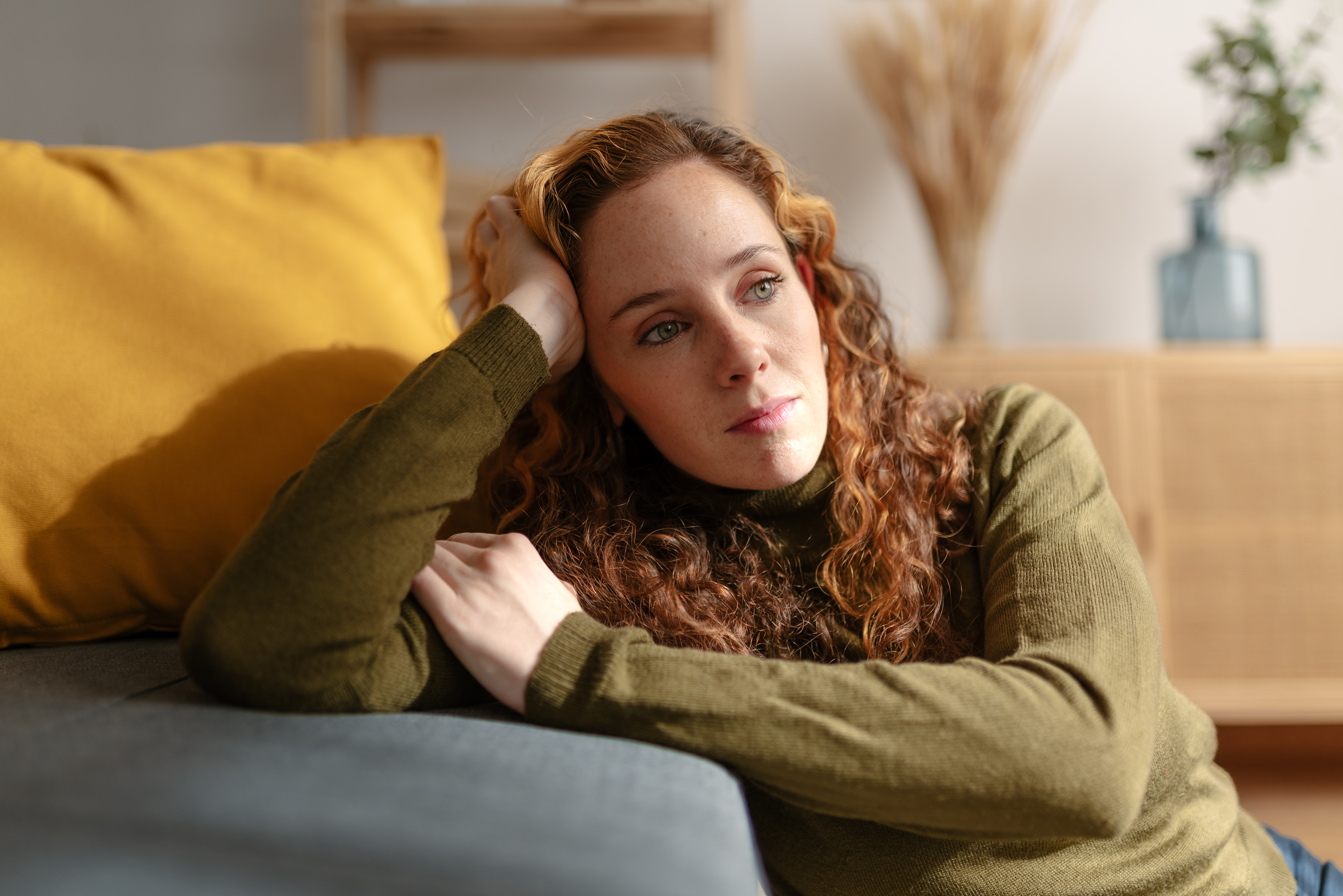 Porträt einer jungen Frau mit langen gelockten roten Haaren, die auf dem Boden sitzt, sich auf ein Sofa stützt und mit traurigem Gesicht wegschaut. Ein Moment der Traurigkeit und Sorge im Wohnzimmer ihres Hauses. | Quelle: Getty Images