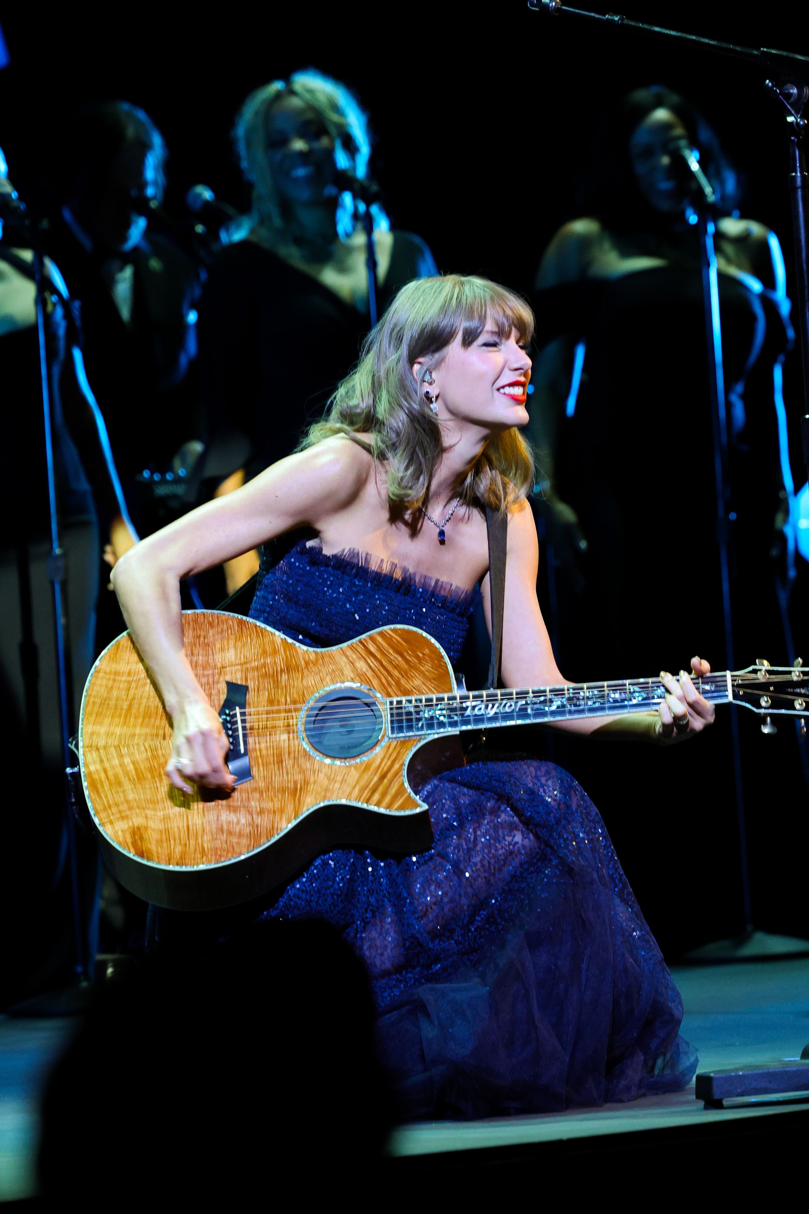 Taylor Swift gibt ein Akustik-Set in einem trägerlosen, mit Pailletten übersäten, mitternachtsblauen Abendkleid, das am Ausschnitt mit zarten Tüllbesätzen verziert ist. Die Sängerin sitzt mit ihrer charakteristischen, maßgefertigten Akustikgitarre da und kombiniert ihren funkelnden Bühnenlook mit einer zarten Saphir-Halskette, Diamant-Ohrringen, ihrem klassischen roten Lippenstift und sanften, honigblonden Locken.