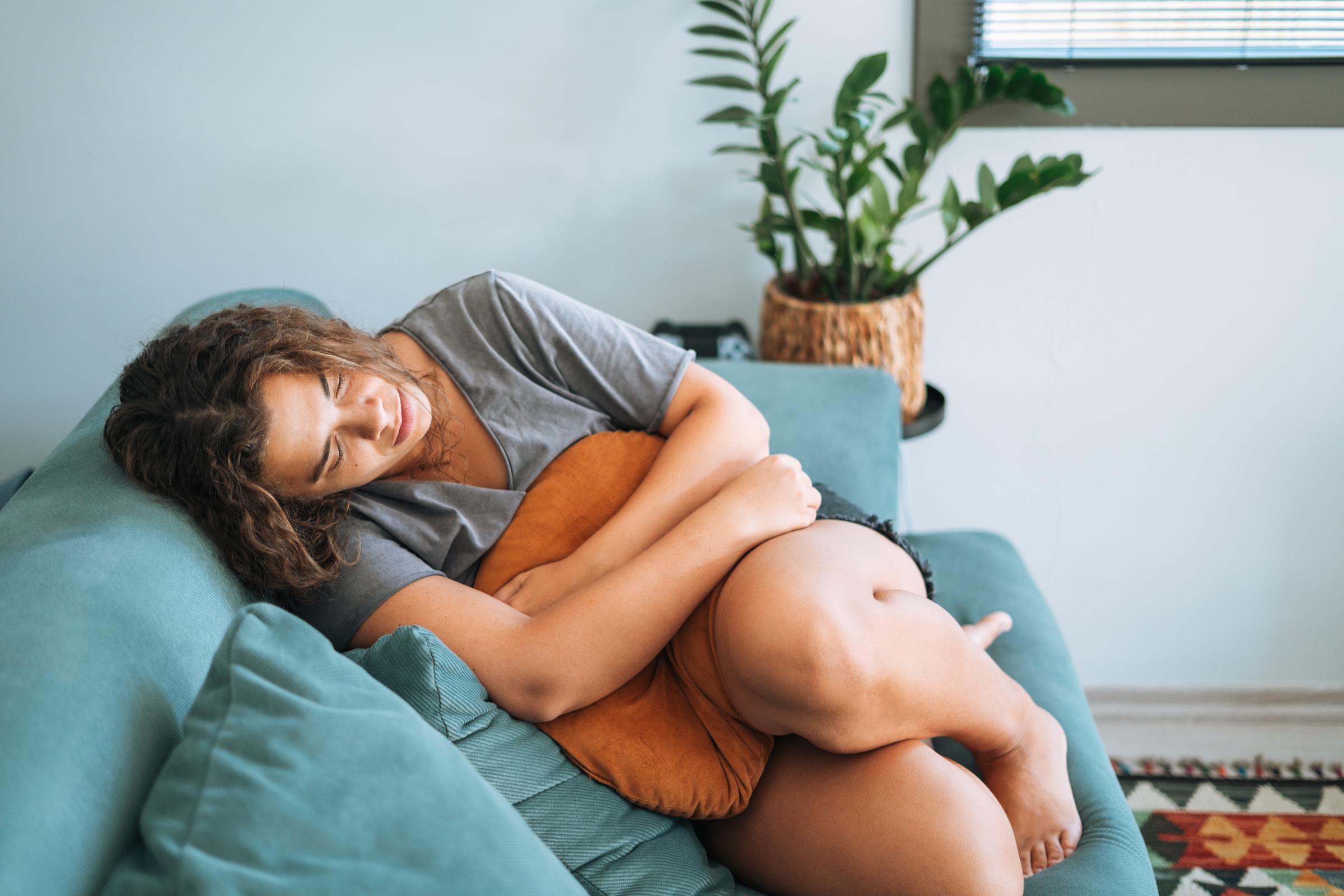 Junge Frau liegt auf einer Couch, hält ein Kissen gegen ihren Bauch und scheint Schmerzen zu haben | Quelle: Getty Images