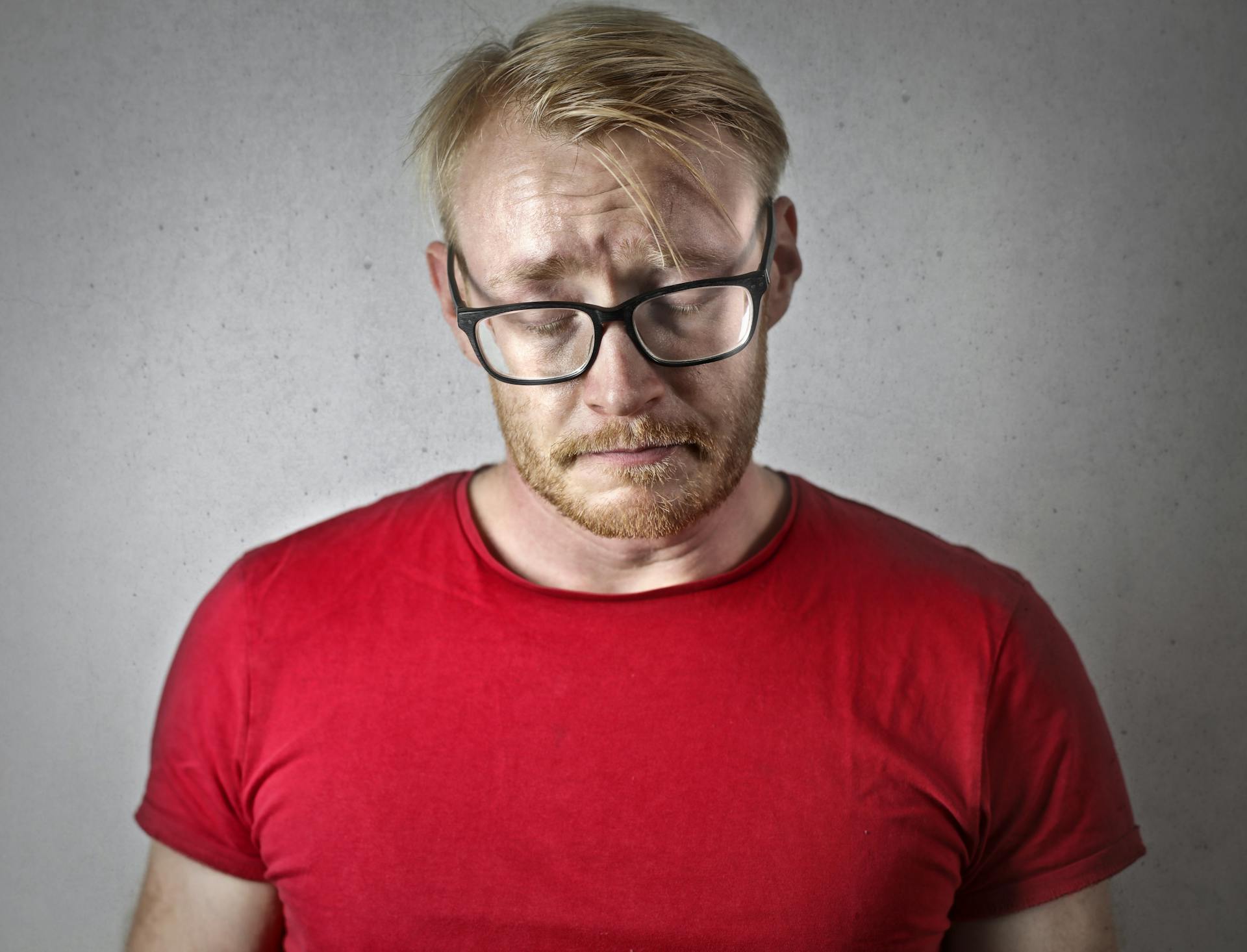 Trauriger Mann in rotem T-Shirt und schwarz gerahmter Brille schließt die Augen | Quelle: Pexels