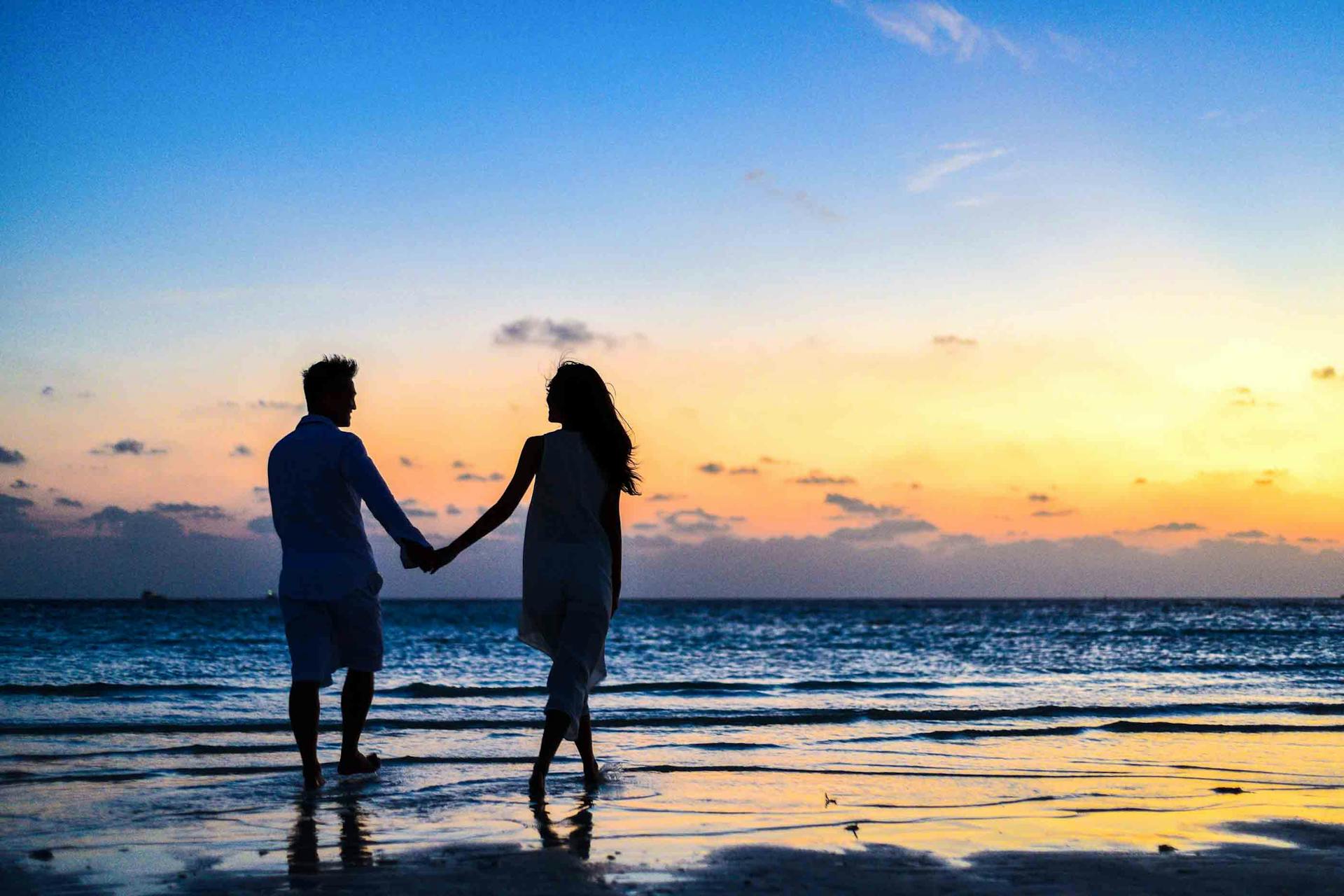 Ein Paar hält sich an den Händen, während es bei Sonnenaufgang am Meer spazieren geht | Quelle: Pexels